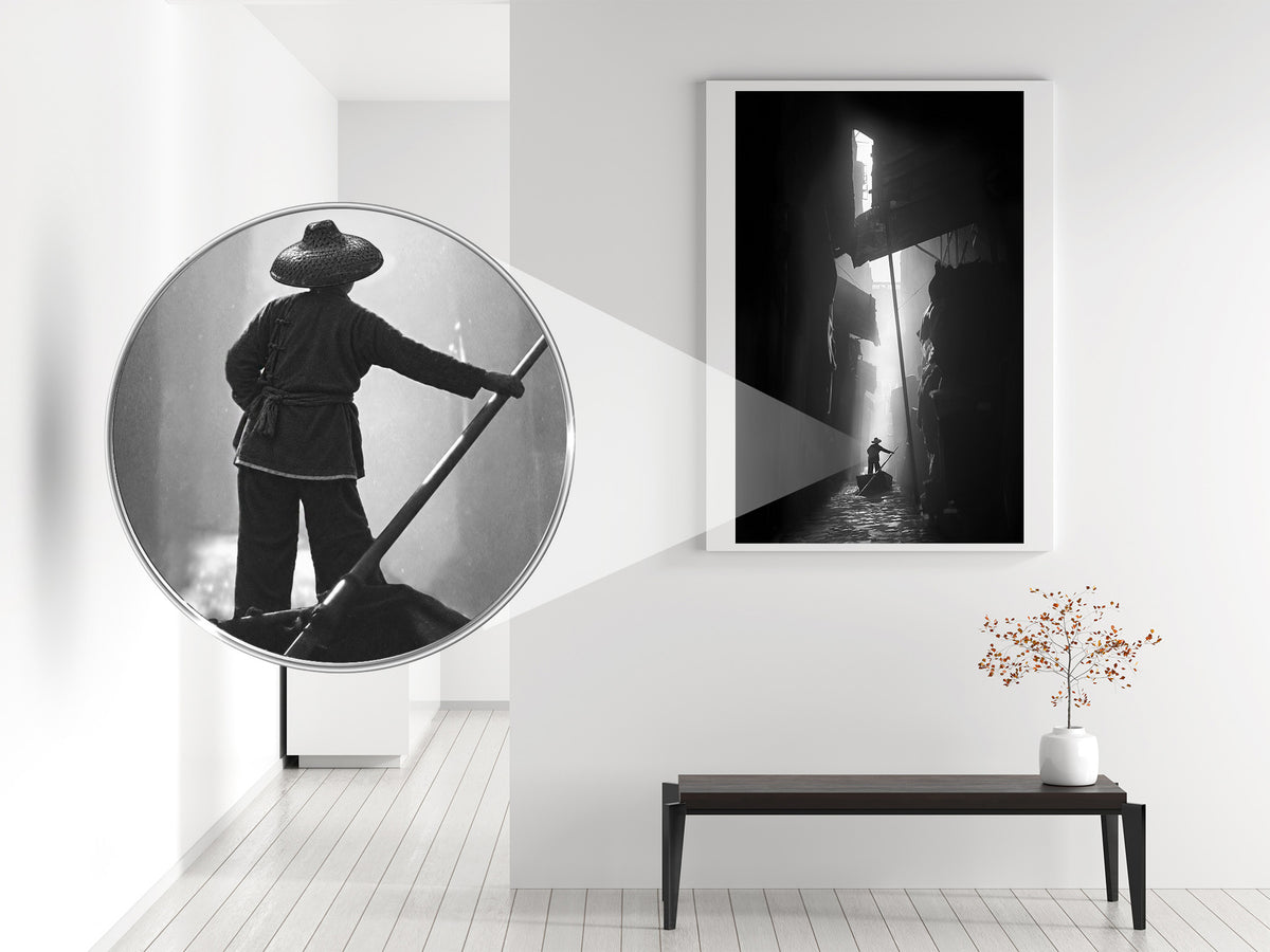 A framed black-and-white wall art print hangs on a white wall in a modern interior, with a bench and vase below. A circular inset shows a close-up of the artwork's subject, a person in a dark outfit holding a long pole in a misty corridor.