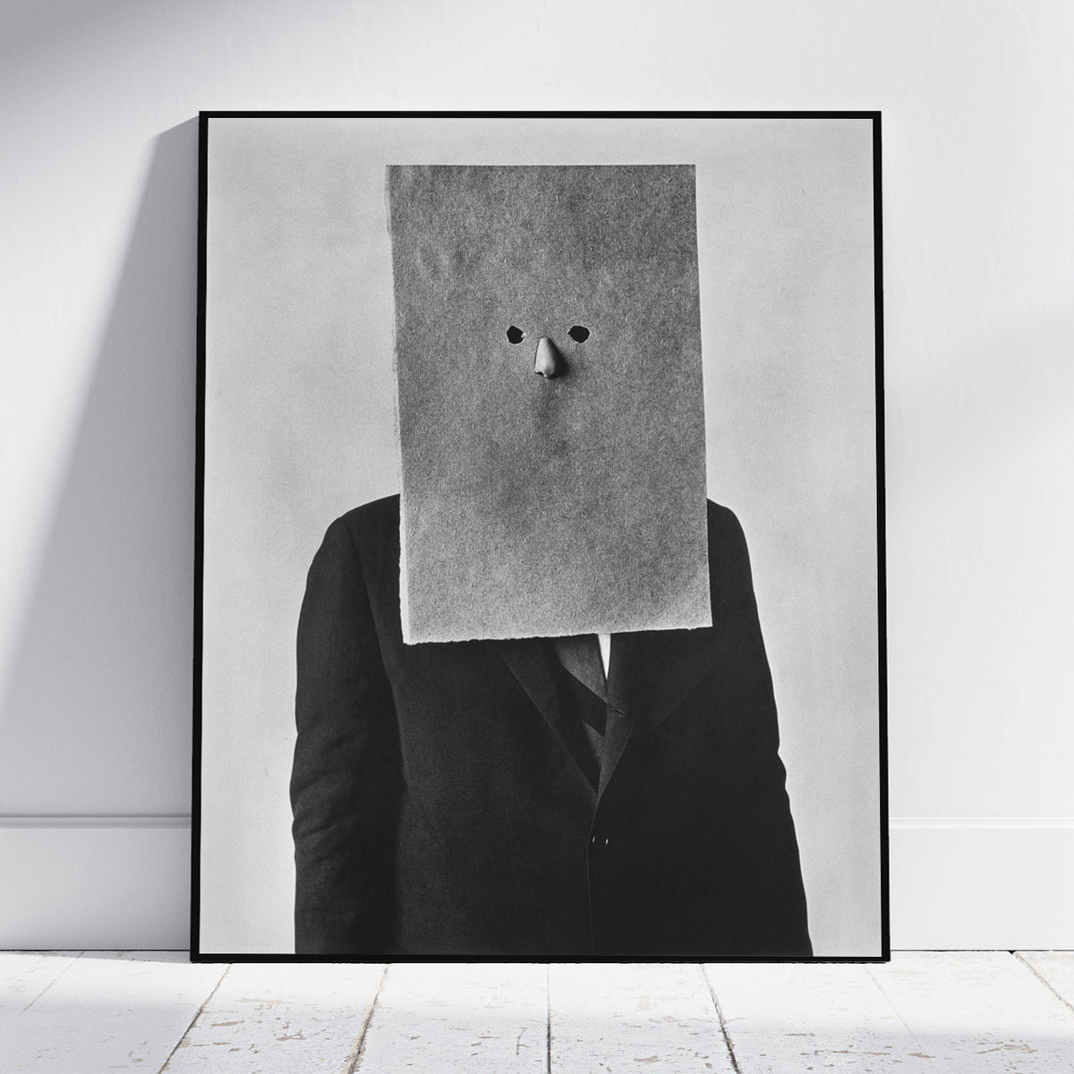 A framed black and white art print leaning against a white wall on a light wood floor. The artwork shows a person in a dark suit with a large rectangular paper bag covering the head, with only small eyes and a nose visible.