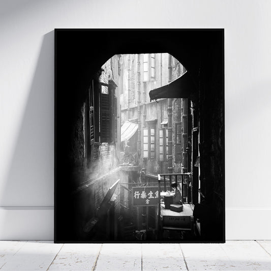 A vertical black and white framed wall art print leaning against a white wall on a light floor. The artwork shows a narrow urban alley with buildings, windows, and street details, framed by a dark arch-like opening.