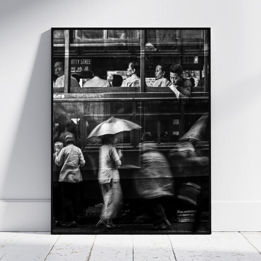 A black and white photographic art print leaning against a white wall on a light floor. The print depicts a city street scene with people, including a figure holding an umbrella, viewed through the windows of a double-decker bus.
