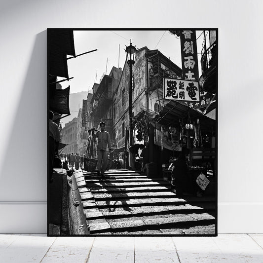 A black and white framed art print depicting a classic Asian street scene with pedestrians, storefronts, and traditional signage, leaning against a white wall on a light wooden floor.
