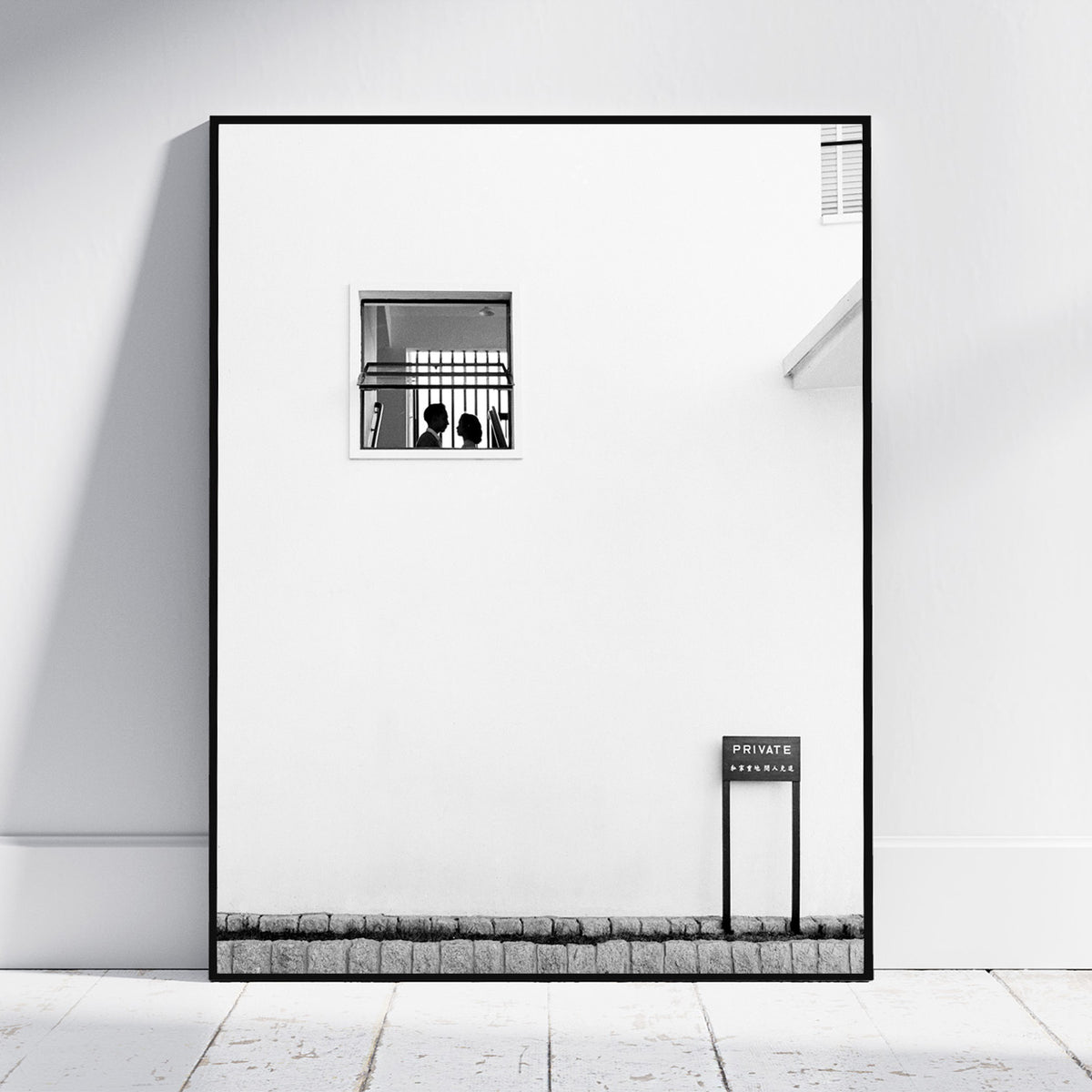 A framed black and white architectural art print leaning against a white wall on a light floor. The print features a minimalist white wall with a small barred window, a small sign reading 'PRIVATE ENTRANCE', and a textured stone-like base.