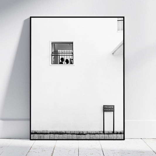 A framed black and white architectural art print leaning against a white wall on a light floor. The print features a minimalist white wall with a small barred window, a small sign reading 'PRIVATE ENTRANCE', and a textured stone-like base.
