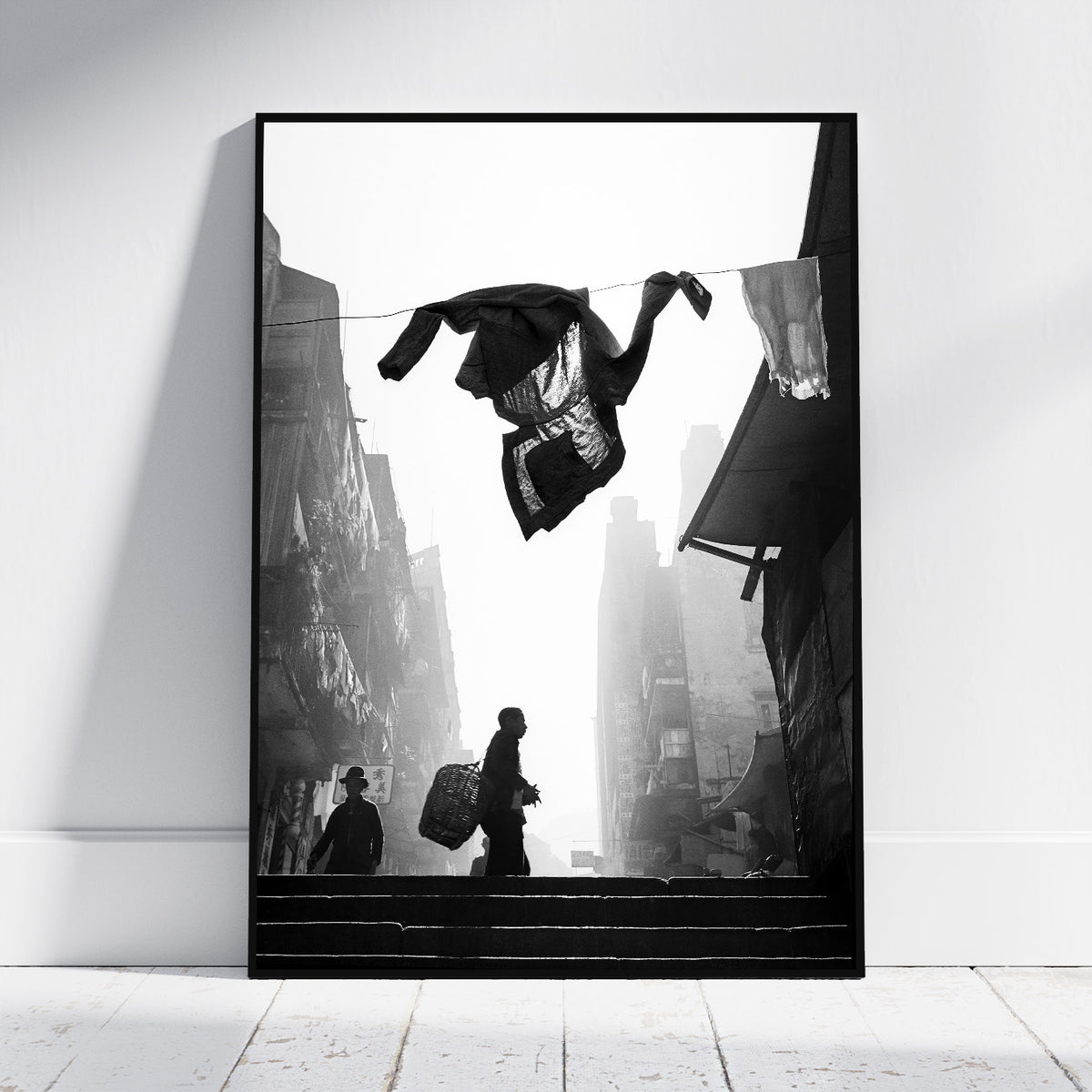 A framed black and white art print leaning against a white wall on a light wood floor. The artwork depicts a street scene with silhouetted figures on stairs and hanging laundry in the foreground.