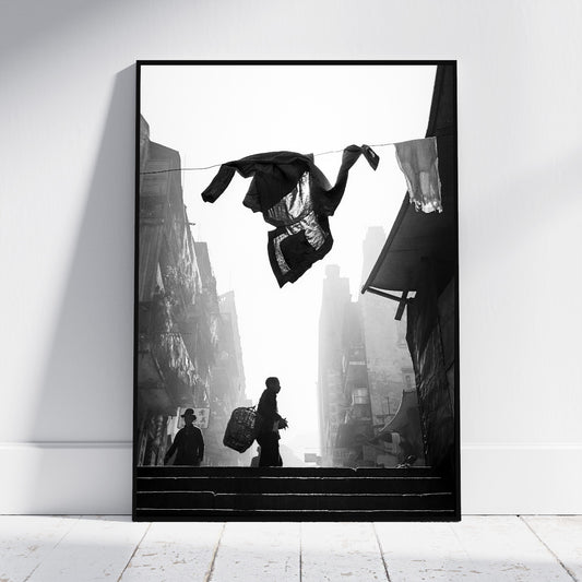 A framed black and white art print leaning against a white wall on a light wood floor. The artwork depicts a street scene with silhouetted figures on stairs and hanging laundry in the foreground.