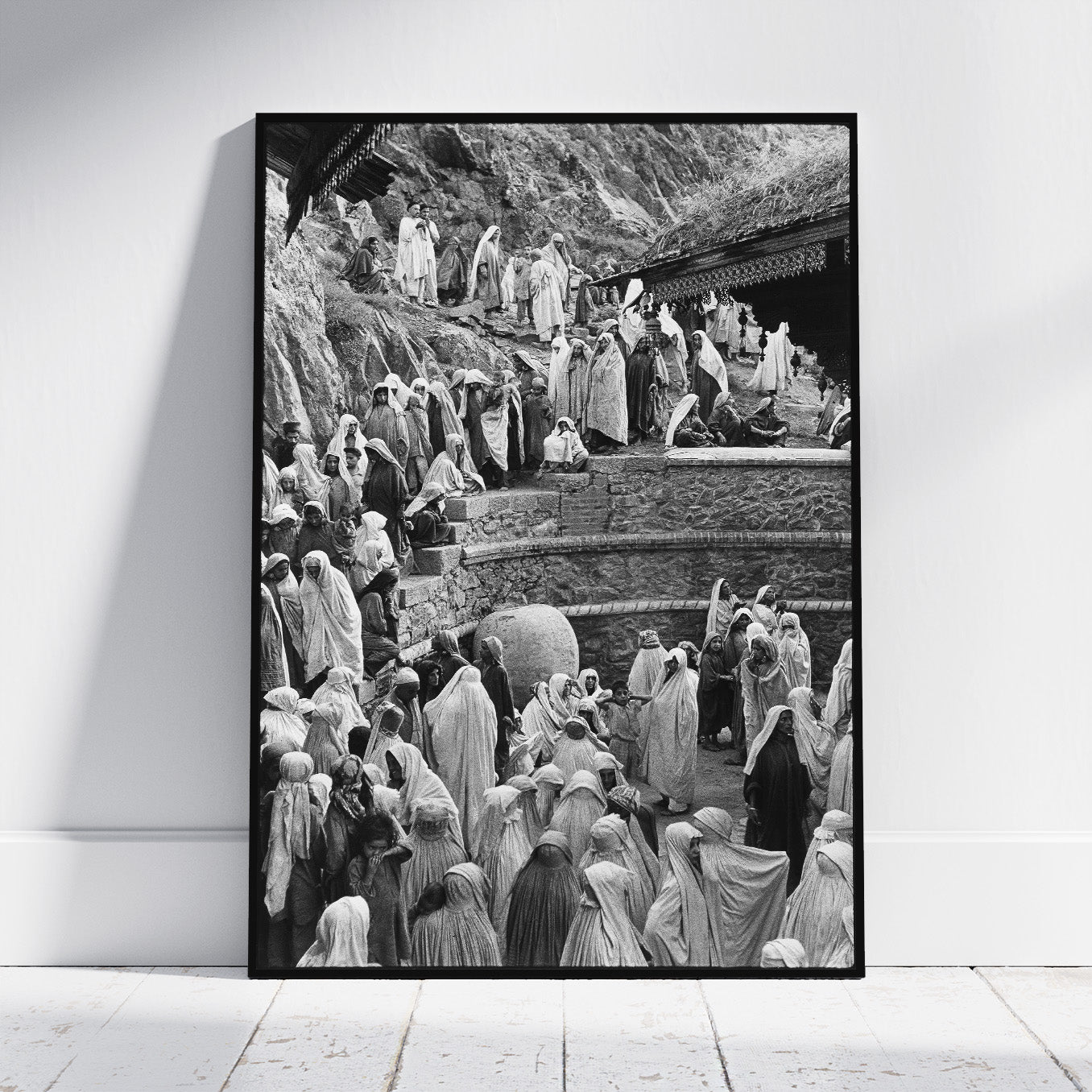 A framed black and white art print leaning against a white wall on a light wooden floor. The print depicts a crowded scene of people in robes on stone steps and a rocky hillside.