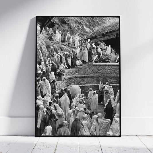 A framed black and white art print leaning against a white wall on a light wooden floor. The print depicts a crowded scene of people in robes on stone steps and a rocky hillside.