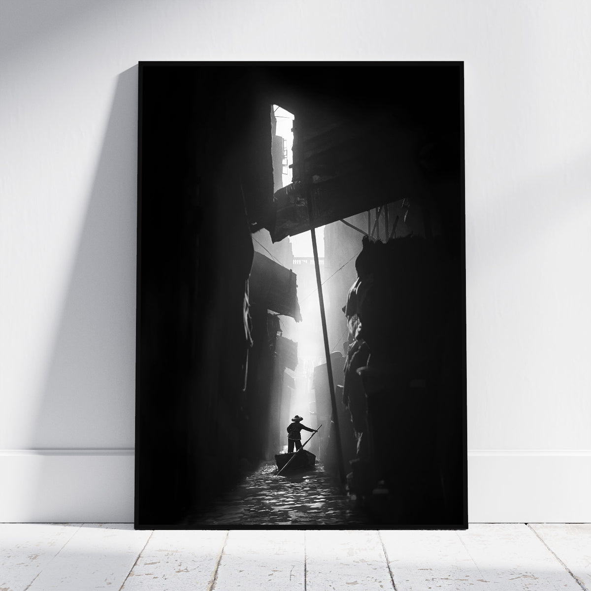 A vertical black and white photographic art print leaning against a white wall on a light wood floor. The print depicts a dramatic scene of a person in a boat navigating a dark, shadow-filled canal with a bright light source at the end.
