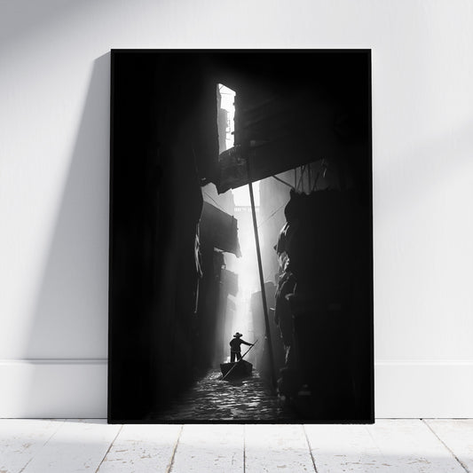 A vertical black and white photographic art print leaning against a white wall on a light wood floor. The print depicts a dramatic scene of a person in a boat navigating a dark, shadow-filled canal with a bright light source at the end.