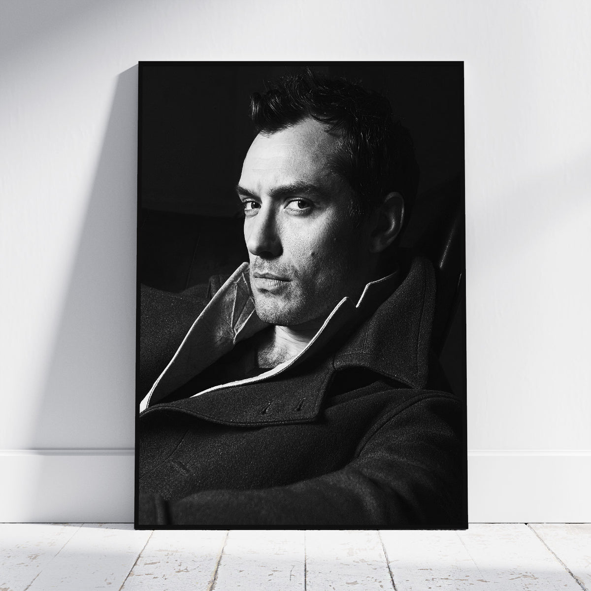A black and white portrait print of a man in a dark coat leaning against a white wall on a light wood floor.