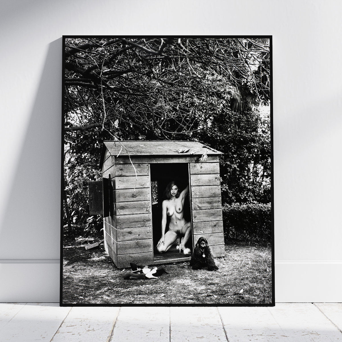 A framed black and white art print leaning against a white wall on a light floor. The print shows a nude woman seated in the doorway of a small wooden shed, with a dog at her side, set against dense trees.