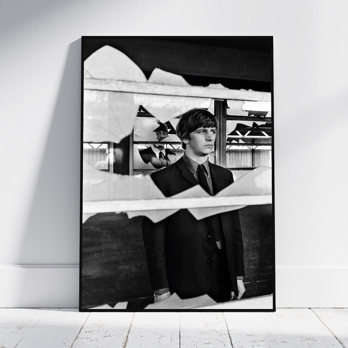A black and white framed art print leaning against a white wall on a light wood floor. The print features a portrait of a young man in a suit seen through a torn-paper effect, with shoes displayed in the background.