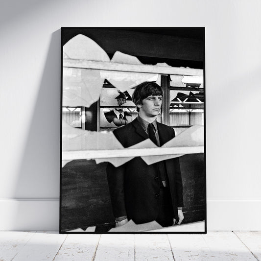 A black and white framed art print leaning against a white wall on a light wood floor. The print features a portrait of a young man in a suit seen through a torn-paper effect, with shoes displayed in the background.
