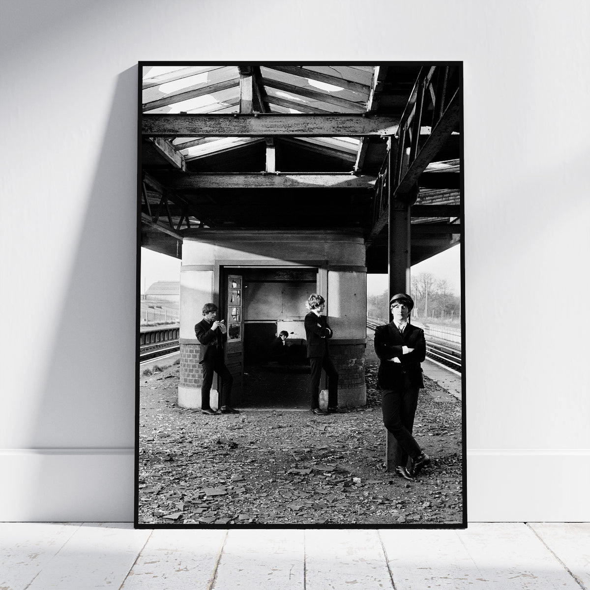 A framed black and white art print leaning against a white wall on a light wooden floor. The print shows three people standing on a train station platform under a bridge.