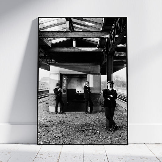 A framed black and white art print leaning against a white wall on a light wooden floor. The print shows three people standing on a train station platform under a bridge.
