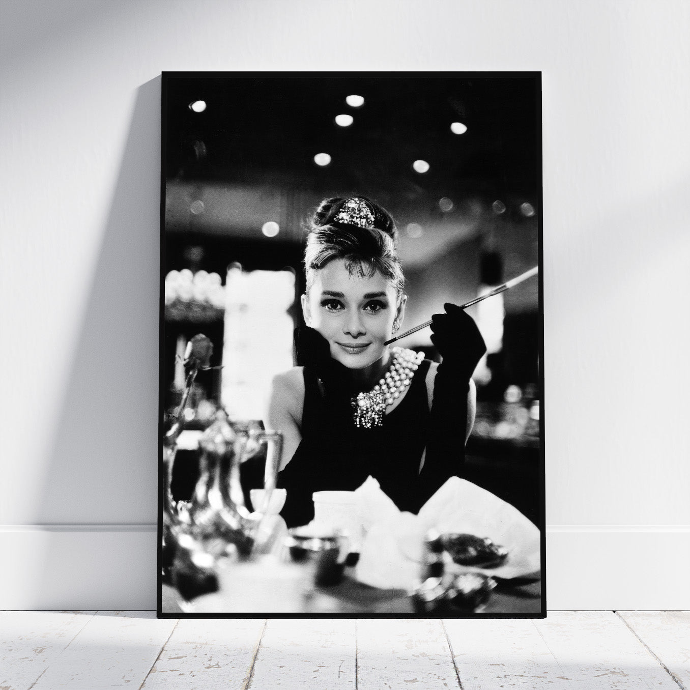 A black and white framed wall art print leaning against a white wall on a light wood floor, showing a monochrome portrait of Audrey Hepburn in a glamorous black outfit with a pearl necklace and cigarette holder.