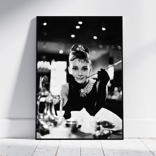 A black and white framed wall art print leaning against a white wall on a light wood floor, showing a monochrome portrait of Audrey Hepburn in a glamorous black outfit with a pearl necklace and cigarette holder.