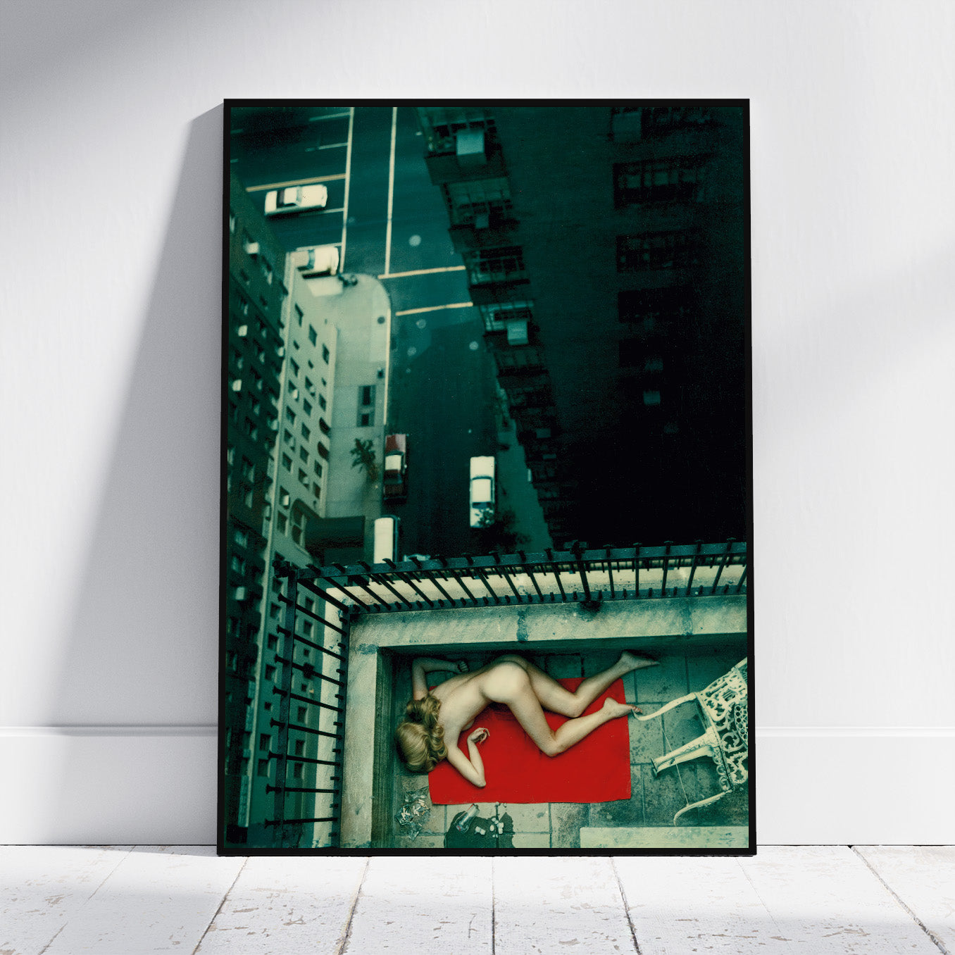 A vertical framed art print leaning against a white wall on a light wood floor. The print shows a high-angle photograph of a nude woman lying on a red mat in an urban courtyard surrounded by tall buildings.