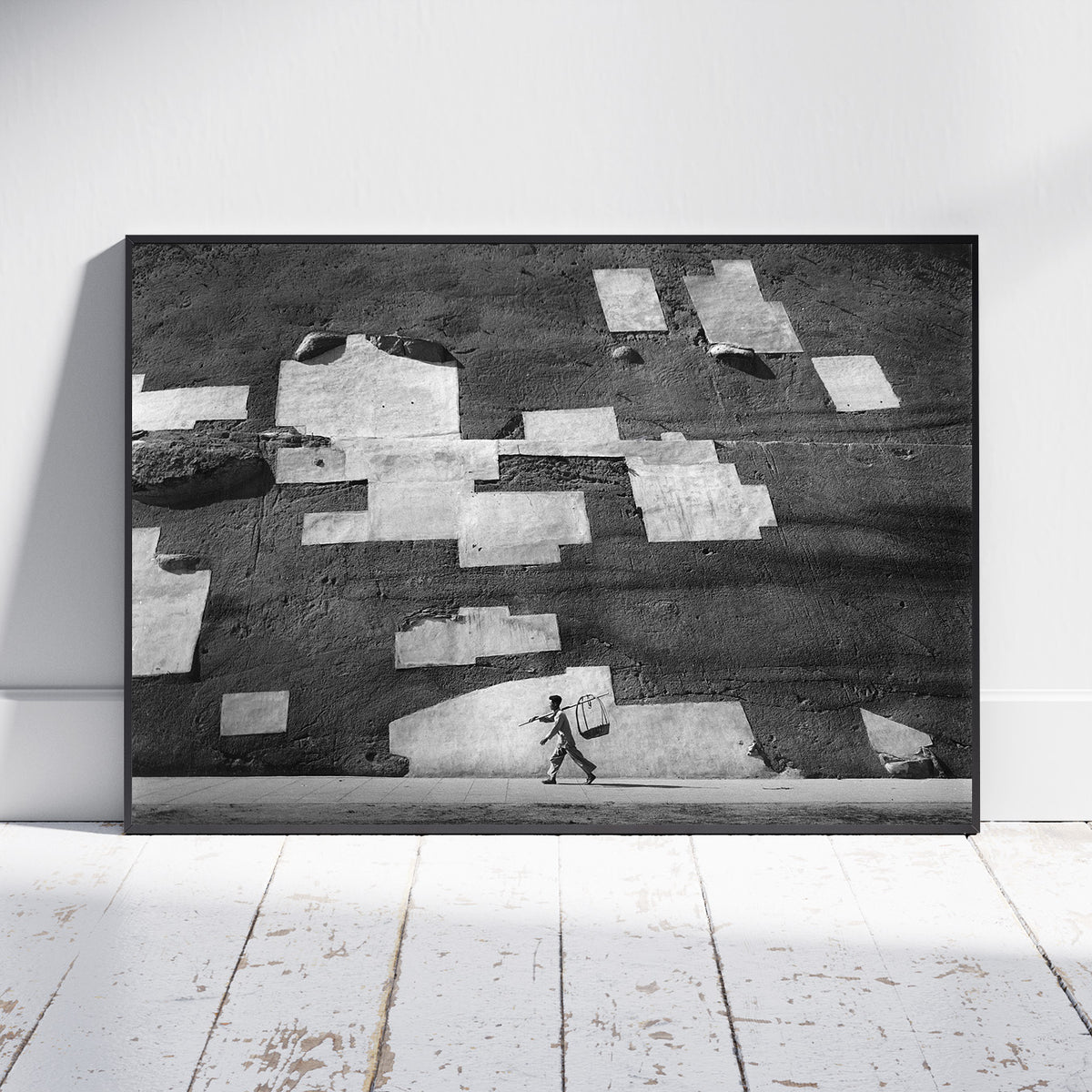 A framed black and white art print leaning against a white wall on a white wooden floor. The artwork depicts a textured wall with irregular light patches and a small walking figure near the bottom.