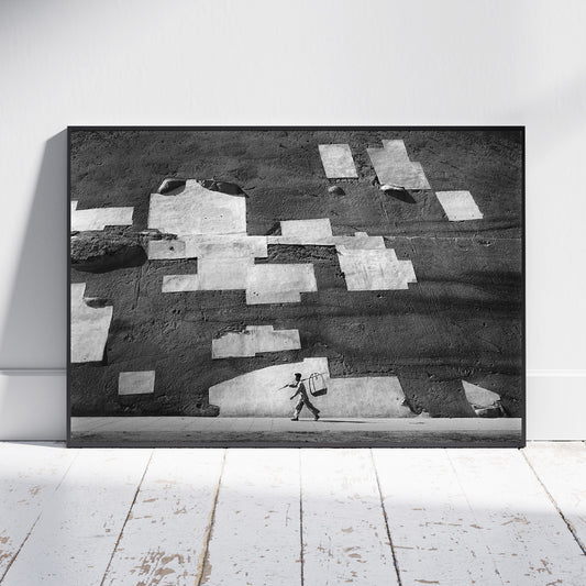 A framed black and white art print leaning against a white wall on a white wooden floor. The artwork depicts a textured wall with irregular light patches and a small walking figure near the bottom.