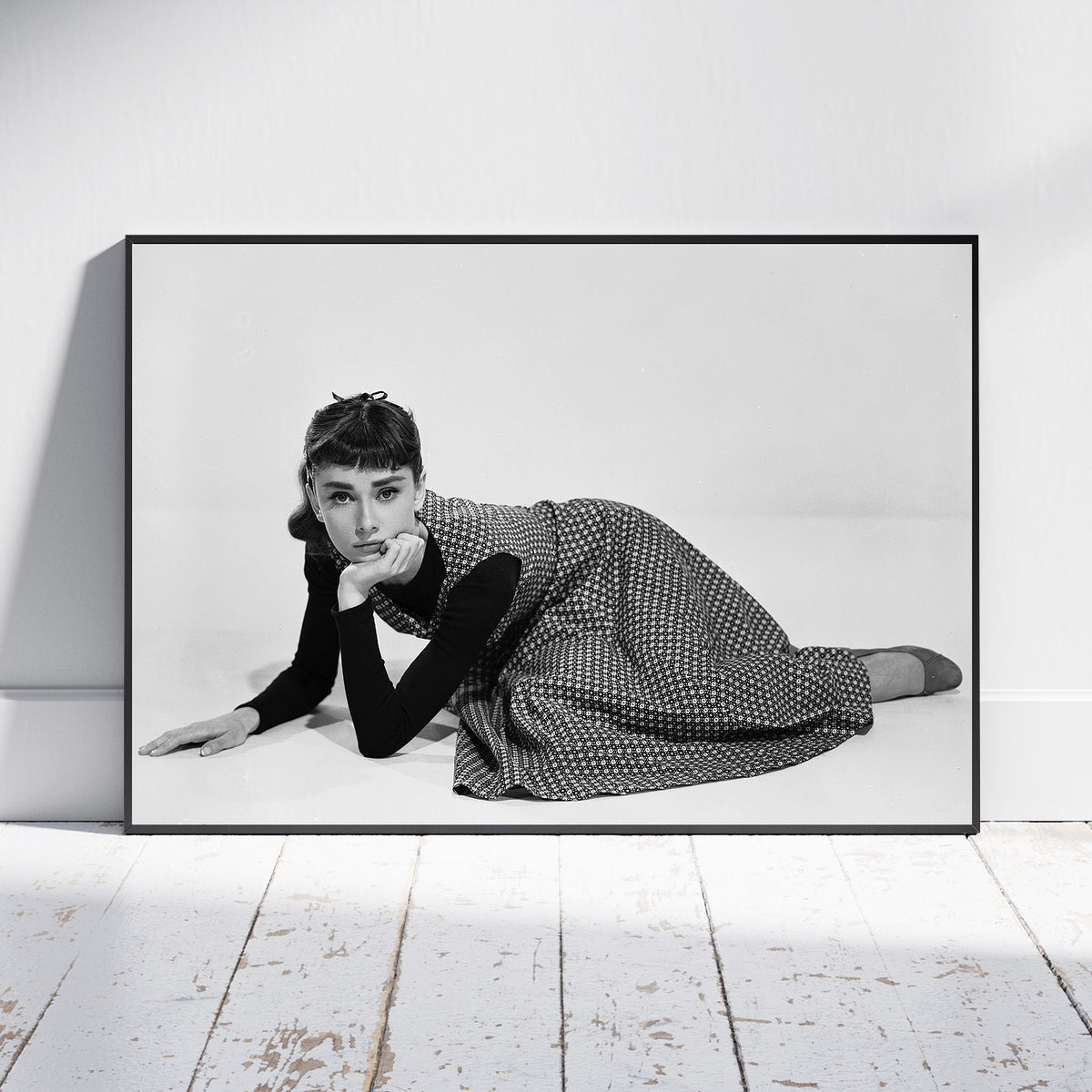 A framed black and white art print of Audrey Hepburn lying on the floor, leaning against a white wall on a light wood floor.