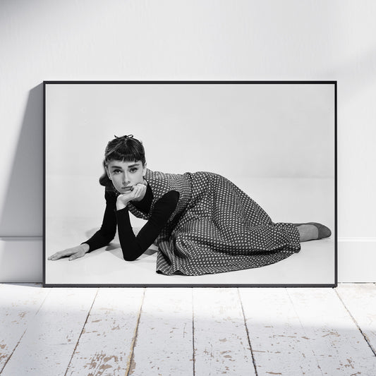 A framed black and white art print of Audrey Hepburn lying on the floor, leaning against a white wall on a light wood floor.