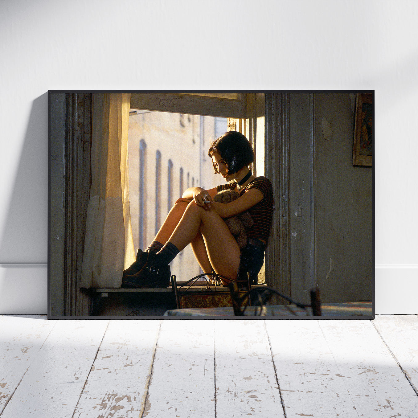 A large horizontal canvas print leaning against a white wall on a white wooden floor. The print shows a woman sitting in a doorway with her legs crossed, wearing a black helmet and boots, with warm sunlight streaming in from outside.