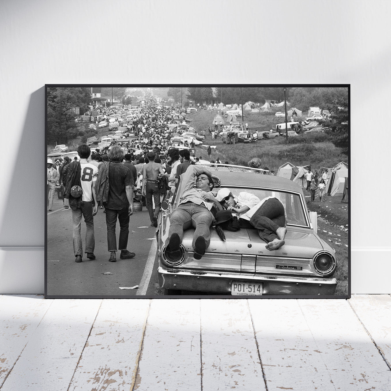 A framed black and white photographic print leaning against a white wall on a white wooden floor. The print shows a crowded outdoor scene with people walking beside and sitting on a vintage car, with tents and vehicles in the background.