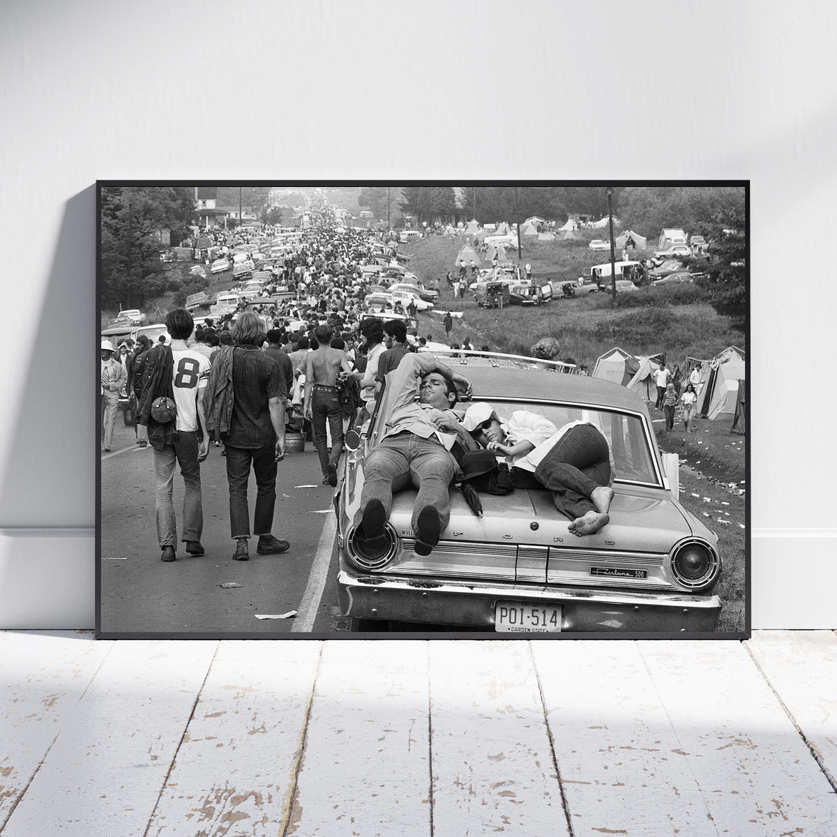 A framed black and white photographic print leaning against a white wall on a white wooden floor. The print shows a crowded outdoor scene with people walking beside and sitting on a vintage car, with tents and vehicles in the background.
