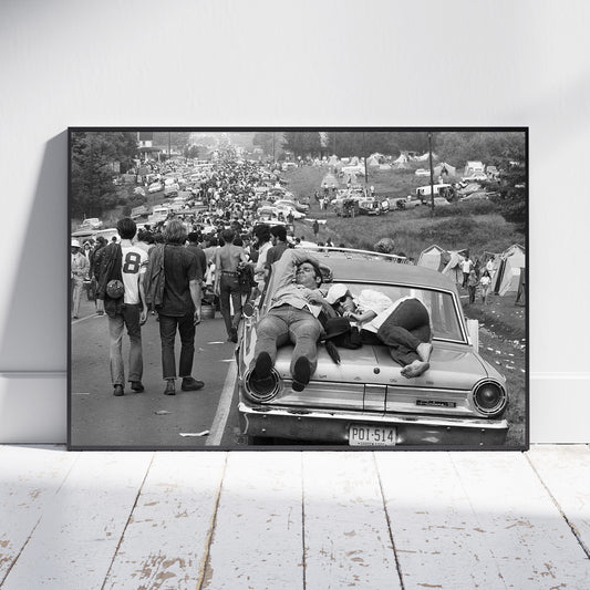 A framed black and white photographic print leaning against a white wall on a white wooden floor. The print shows a crowded outdoor scene with people walking beside and sitting on a vintage car, with tents and vehicles in the background.
