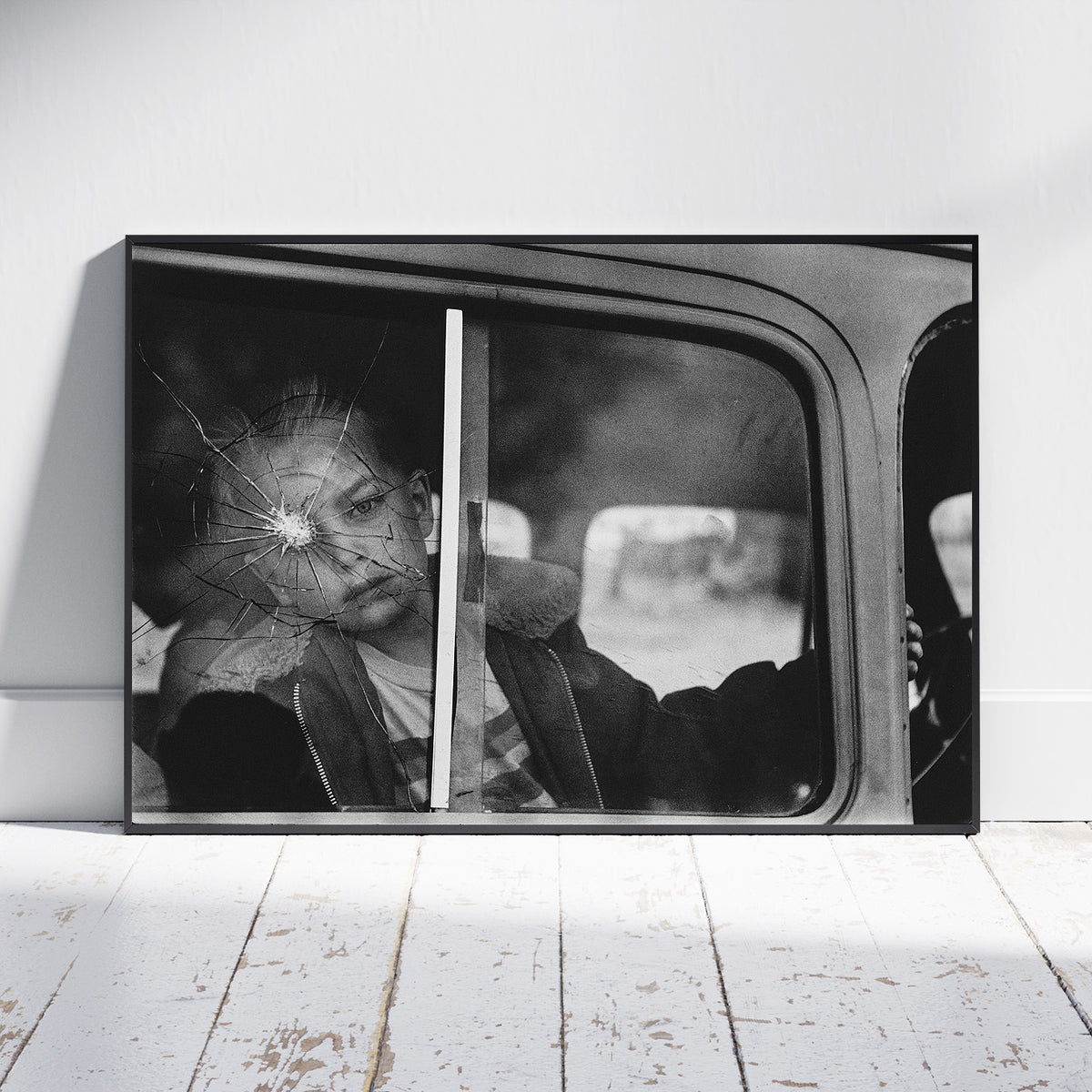 A framed black and white photographic print leaning against a white wall on a white wooden floor. The artwork shows a person sitting inside a vehicle, seen through a cracked window.