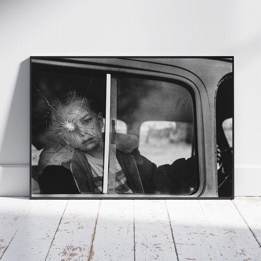 A framed black and white photographic print leaning against a white wall on a white wooden floor. The artwork shows a person sitting inside a vehicle, seen through a cracked window.