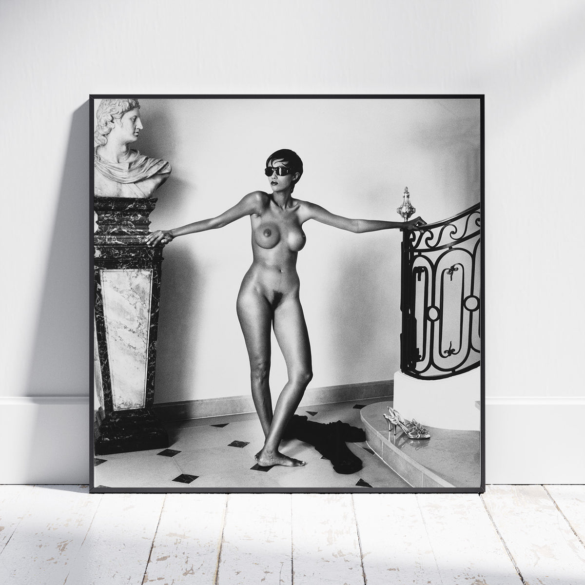 A framed black and white art print leaning against a white wall on a light wood floor. The print shows a nude woman standing indoors with arms outstretched beside a marble bust and an ornate railing.