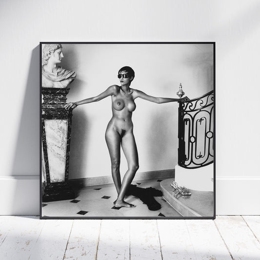 A framed black and white art print leaning against a white wall on a light wood floor. The print shows a nude woman standing indoors with arms outstretched beside a marble bust and an ornate railing.