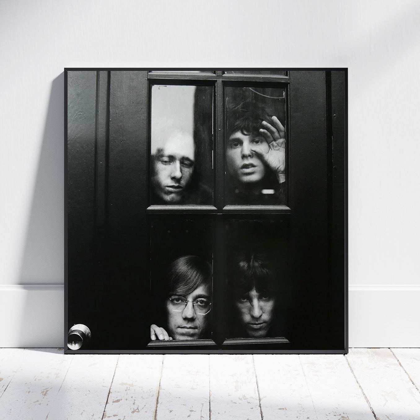 A square black-framed wall art print leaning against a white wall on a white wood floor. The artwork shows a black-and-white four-pane window-style composition with four faces looking in.